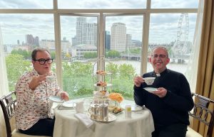Alan and Richard having high tea in a hotel. Big windows showing the banks of the River Thames. London Eye in the background.