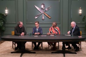 Richard and three other people sat at a large oval table. Dark green panelled wall with a large knife, fork and spoon logo on the wall.