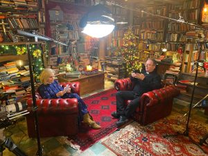 Sally and Richard on set. Sat on red Chesterfield seats in a room with book shelves on all the walls. Christmas tree in the corner. Light and sound stands visible.