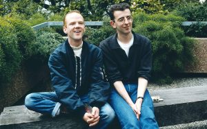 Communards sat on a wooden bench in a garden. Pack of Marlboro lights to the side.